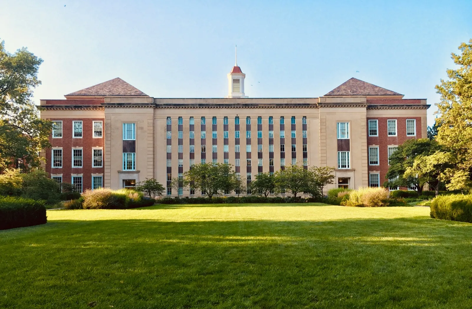 A brick college campus building with columns and a green lawn on a sunny day