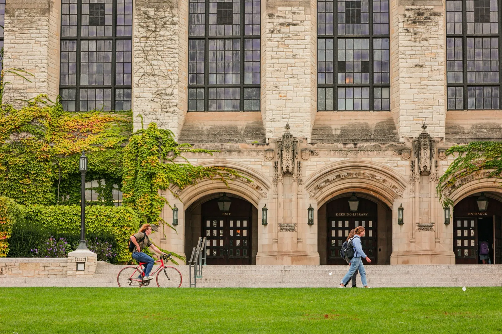 An ivy-covered stone campus building with arched entrances and students walking on the green lawn