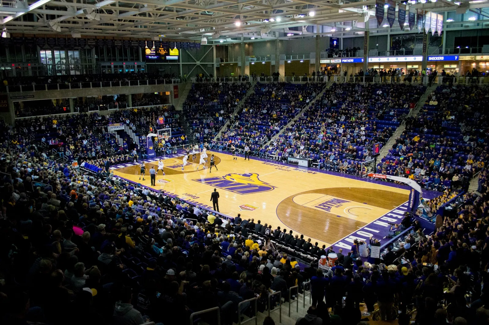 A packed college basketball arena during a game, with fans filling the stands and players on the hardwood court