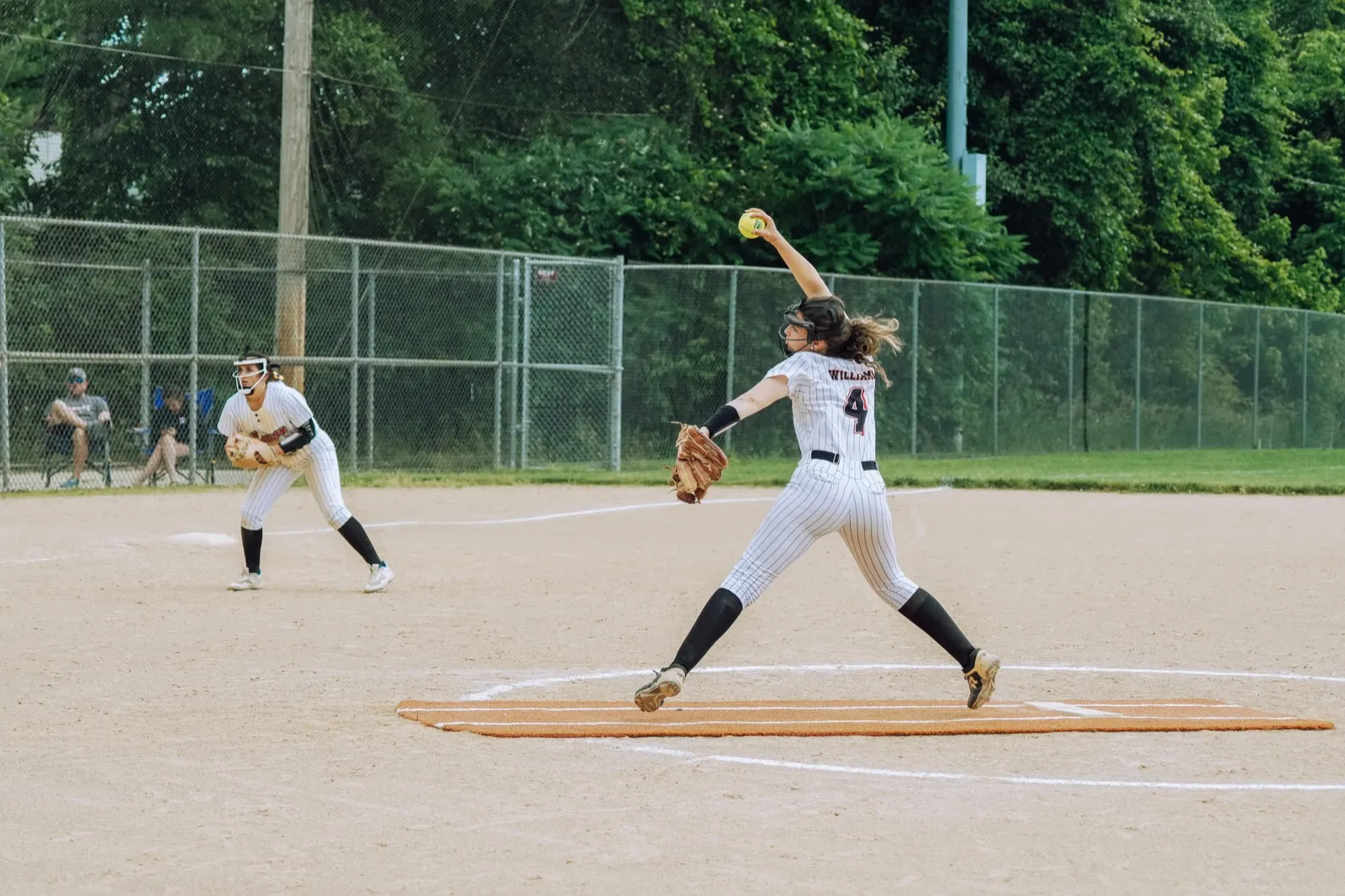 A softball pitcher in a white uniform in her windup on the mound