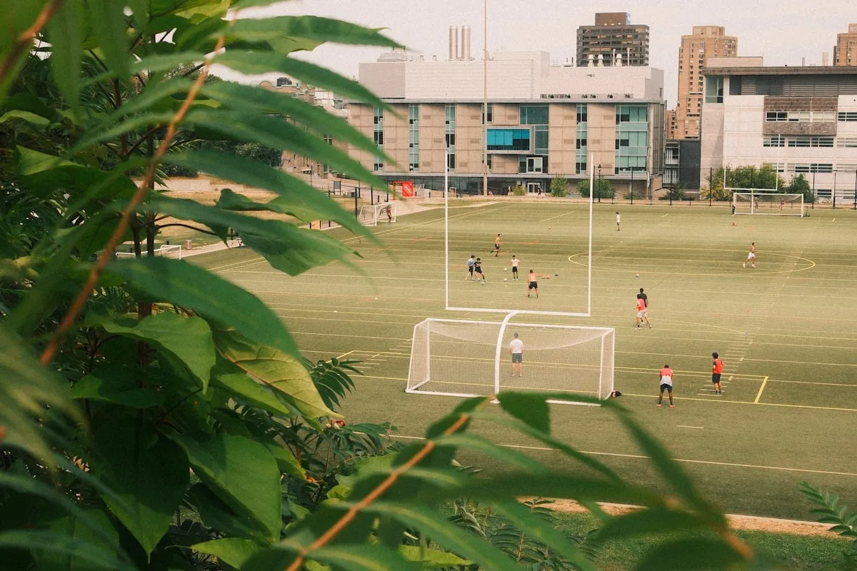 Wide view of a college soccer field with campus buildings in the background and players on the pitch
